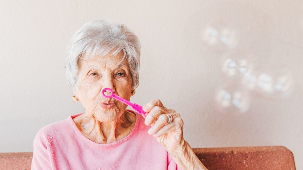 A frail woman with white hair and a pink top playfully blows bubbles through a pipe cleaner bubble wand